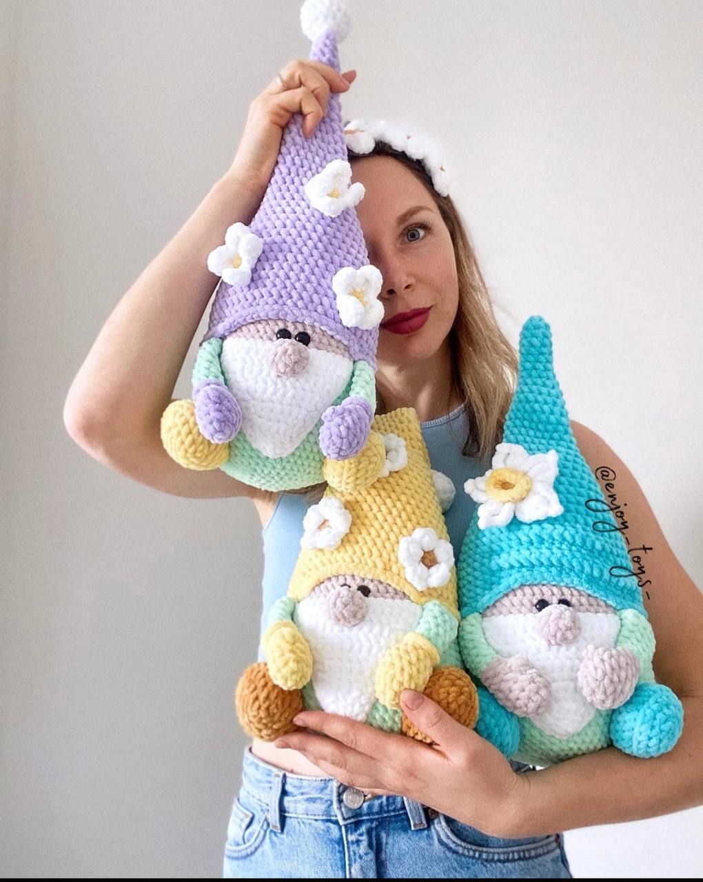 Handmade pastel crochet gnomes with decorative flowers, held by a woman wearing a daisy headband. Artisan amigurumi dolls woven with soft yarn.