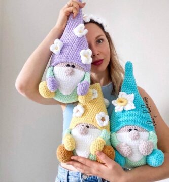 Handmade pastel crochet gnomes with decorative flowers, held by a woman wearing a daisy headband. Artisan amigurumi dolls woven with soft yarn.